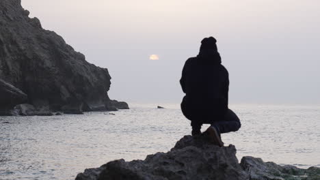 man jumps onto a rough rock and sits down to watch sunrise as seagulls fly over sea, fascinated by beautiful view