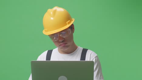 close up de hombre asiático trabajador con gafas de protección y casco de seguridad usando una computadora portátil y con dolor de cabeza mientras está de pie en el estudio de fondo de pantalla verde