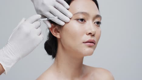 hands in gloves giving injection in cheek of asian woman on grey background, slow motion