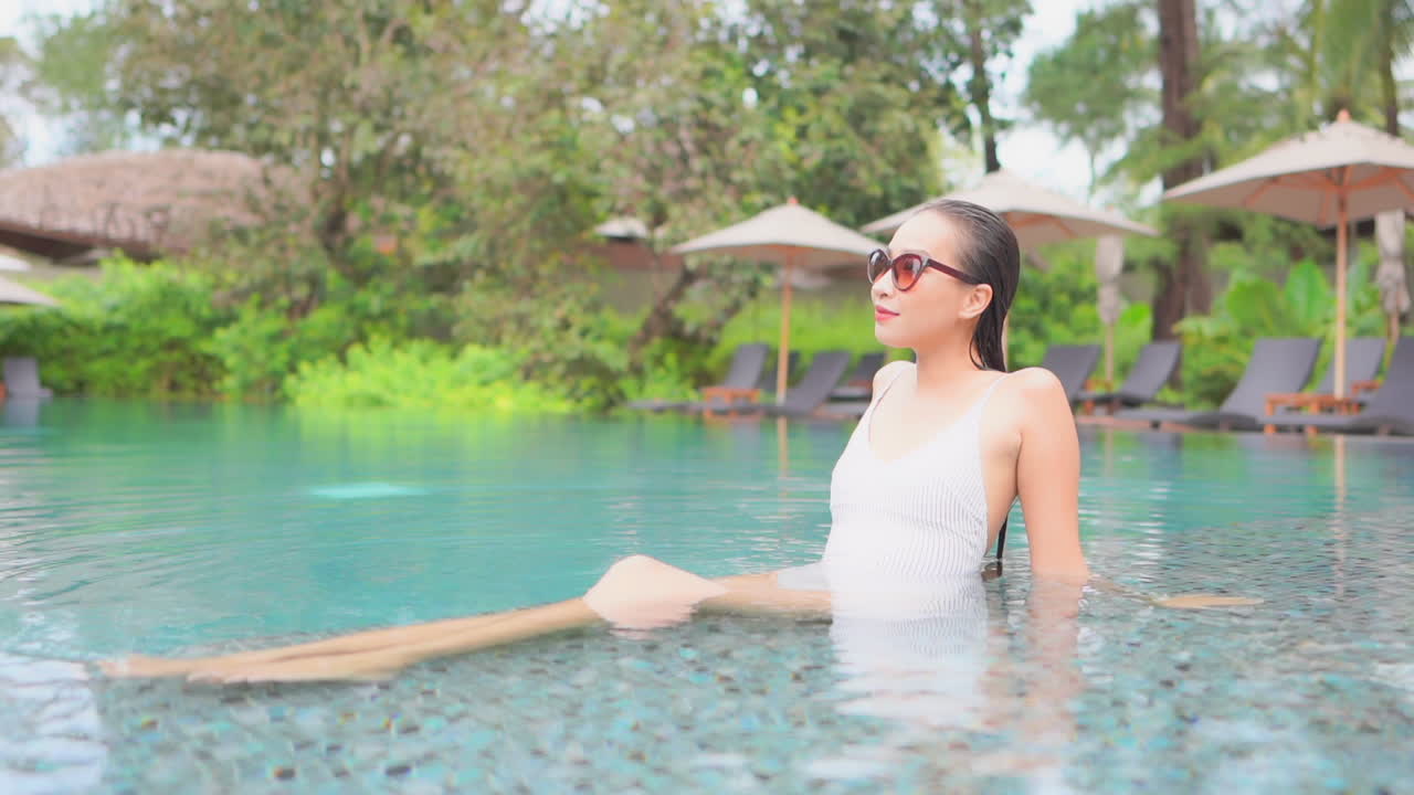 이국적인 인도네시아 호텔 라운지의 얕은 수영장 물에서 선글라스와 흰색 수영복을 입은 똑똑한 여성, 배경에 파라솔과 데크 의자 줄