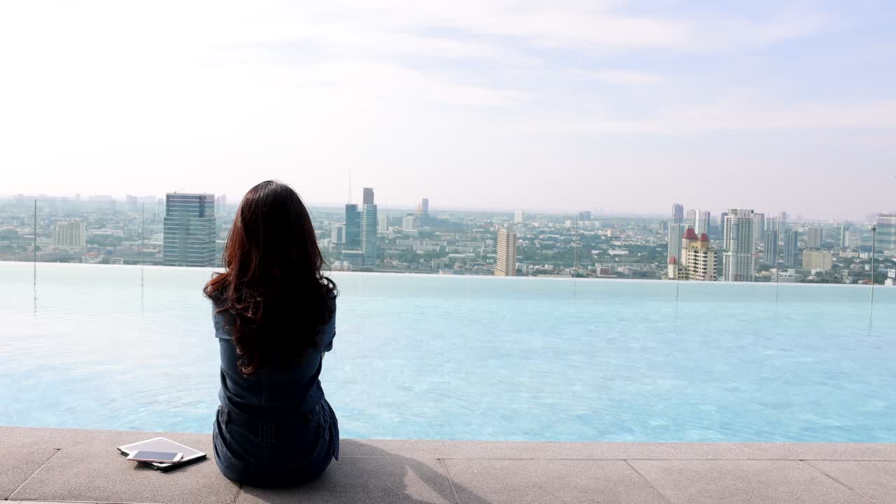 vista trasera de una mujer sentada en el borde colgando sus pies en la piscina en un rascacielos, usando la red social en un teléfono inteligente, tableta. fondo borroso ciudad moderna y hermoso cielo, nubes. espacio de copia