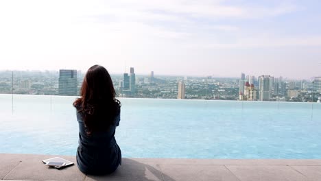 back view of woman sitting on the edge dangling her feet in swimming pool on skyscraper, using social network on smartphone, tablet. blurred background modern city and beautiful sky,clouds. copy space