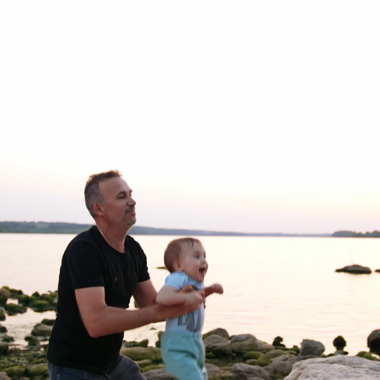 Tempo in famiglia nella natura. Un padre gioca con il suo bambino vicino alla riva del fiume. Superficie dell'acqua calma sullo sfondo