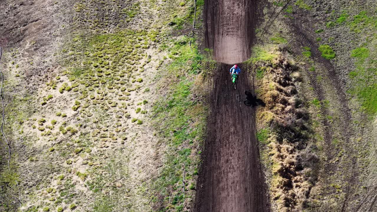 A motorbike rider launches off a dirt jump on a rural track, captured in bright daylight by a stationary aerial drone in Tregony, Queensland