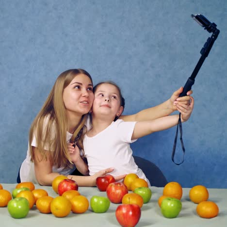 beautiful woman and little girl doing selfie. happy mother and daughter doing photo on a phone near the table with fresh fruit indoors.