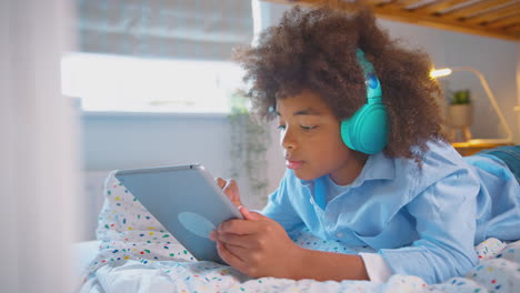 niño en el dormitorio acostado en la cama usando auriculares usando tableta digital