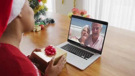 Mujer-Afroamericana-Con-Gorro-De-Papá-Noel-Usando-Una-Computadora-Portátil-Para-Una-Videollamada-Navideña,-Con-La-Familia-En-La-Pantalla