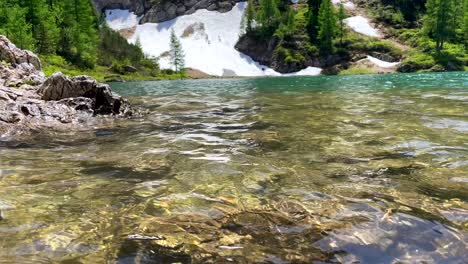 Low-angle-shot-of-clear-fresh-mountain-lake-in-sunlight-and-snowy-shore-in-background