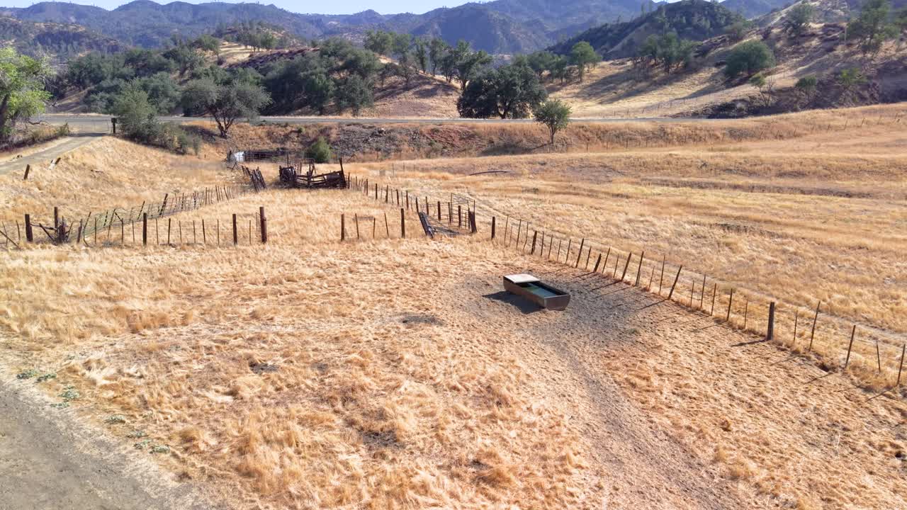 An aerial view of a rustic California ranch, showing a water trough and wooden fence in a dry, golden pasture. A classic scene of rural life in the American West