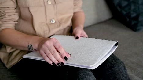 the camera focuses on woman hand reading a braille book sitting on the sofa at home 1