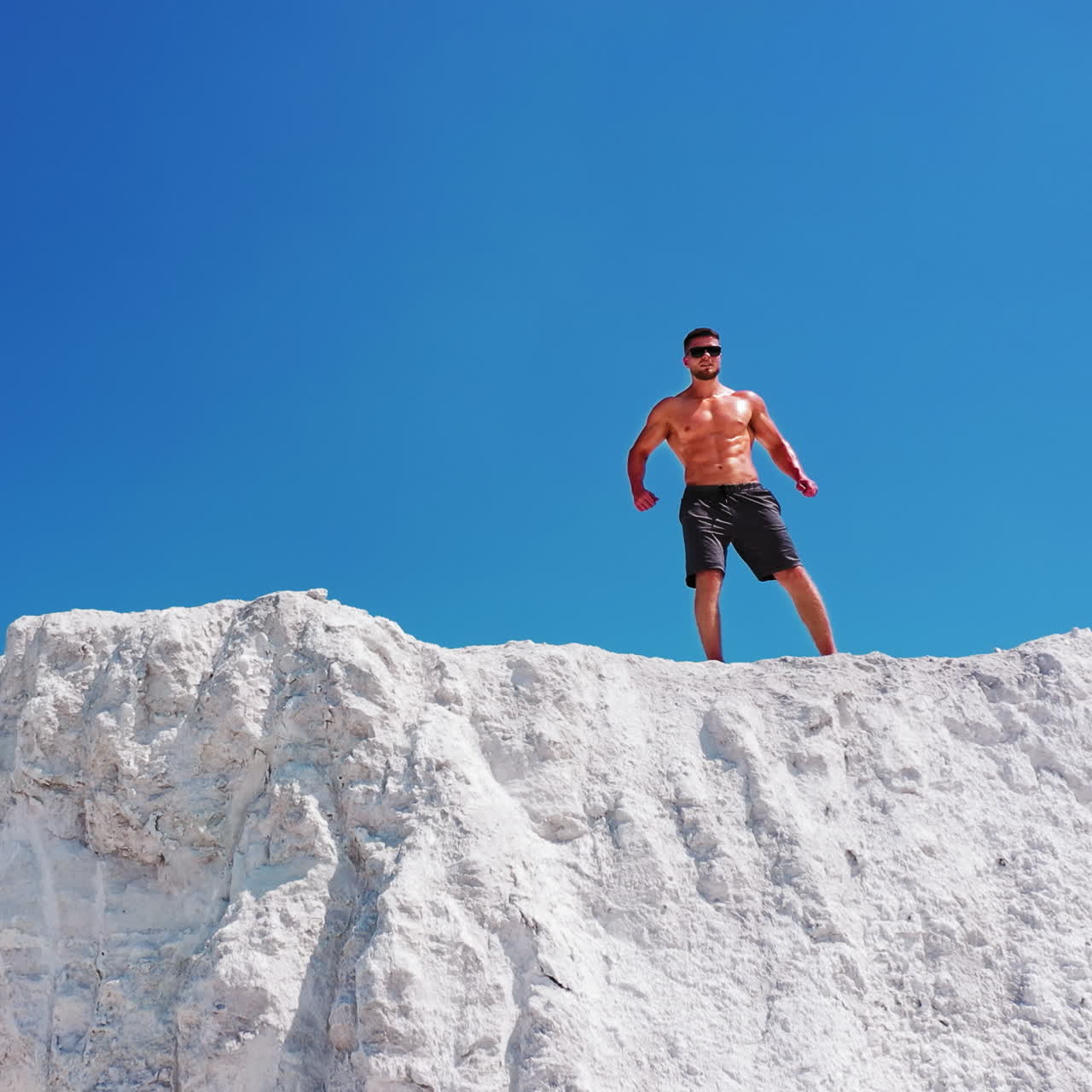 Muscular sportsman under blue sky. Strong athlete in black sunglasses posing on camera on the top of white mountain. Camera moves forward. Drone view.