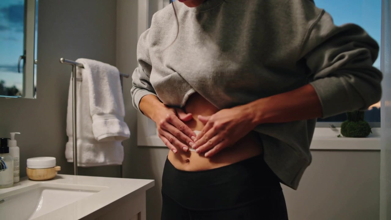 Donna con top grigio che allunga la mano, stacca la pellicola protettiva e applica un cerotto sullo stomaco in bagno per sollievo.