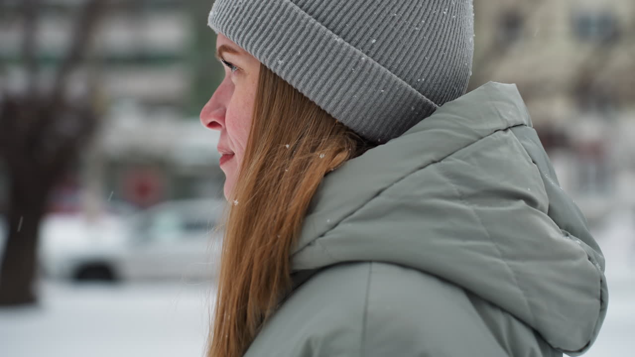 Young woman in gray beanie and winter coat looks down peacefully in snowy urban setting, soft snowflakes on hat and hair, blurred cars and buildings in background, tranquil moment during cold weather