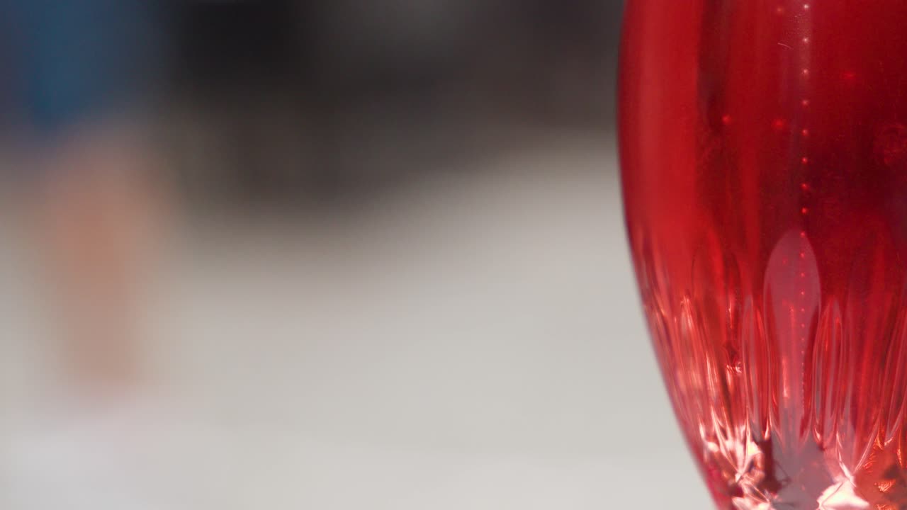 Crystal glass with red drink in sharp focus, blurred people and soft evening outdoor lighting
