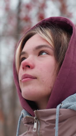 primer plano de una mujer con una chaqueta de invierno con capucha, sonriendo cálidamente mientras camina a través de un ambiente sereno y cubierto de nieve, un fondo borroso con árboles desnudos y bayas rojas
