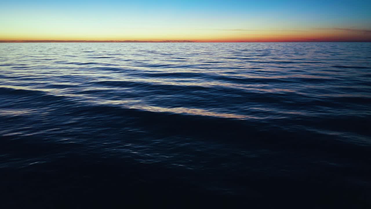 Ocean waves gently ripple on the surface as the sun sets below the horizon, casting warm, muted colors across the vast, tranquil sea under a clear sky