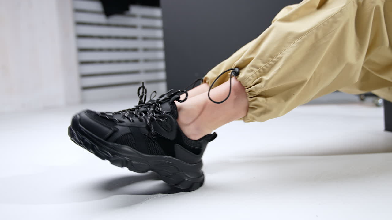 Black trendy massive sneakers on female feet. Woman wearing beige broad pants sits on the floor demonstrating her sport shoes.
