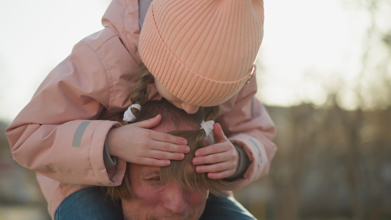 Playful father-daughter moment in a park, as she sits on his shoulders, covers his eyes, and smiles, both enjoying the sunny day