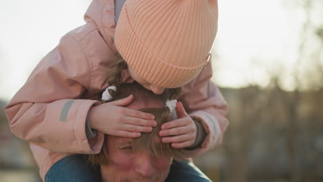 momento juguetón padre-hija en un parque, mientras ella se sienta en sus hombros, cubre sus ojos, y sonríe, ambos disfrutando del día soleado