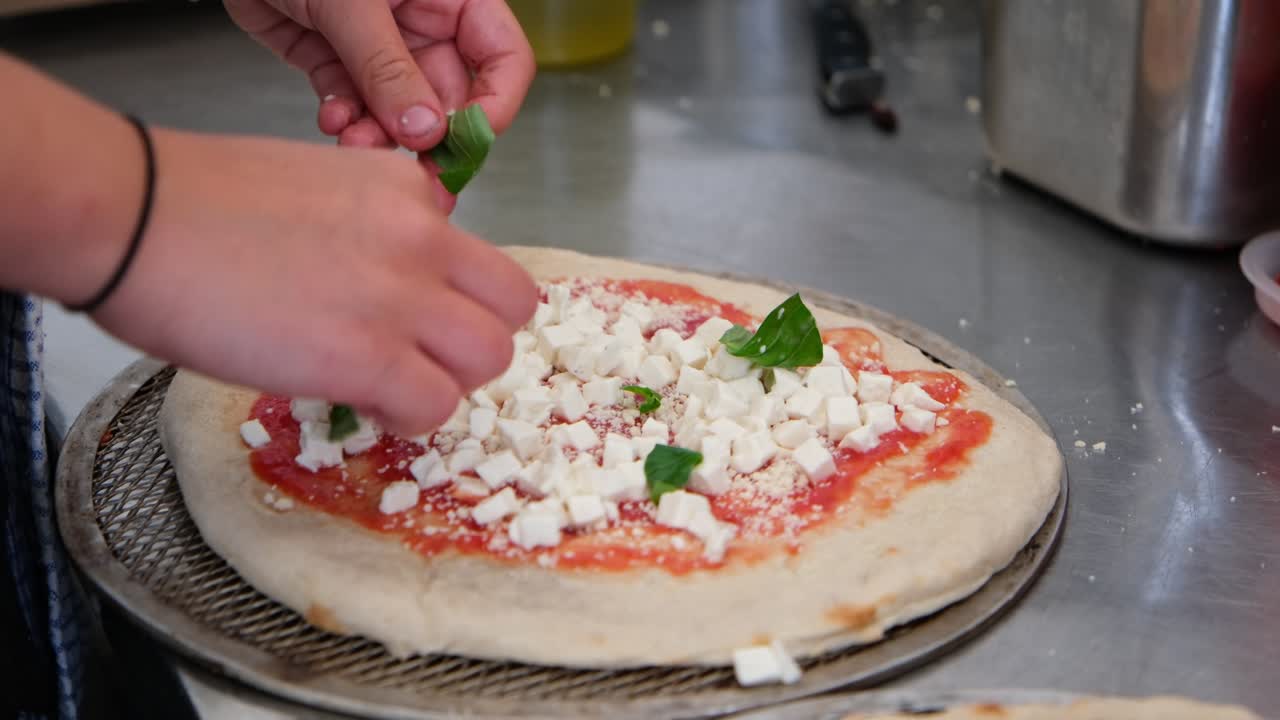 Close-up of hands in pizzeria topping pizza with fresh basil leaves and drizzling olive oil over cheese and tomato sauce before baking. Authentic Italian cooking and preparation process