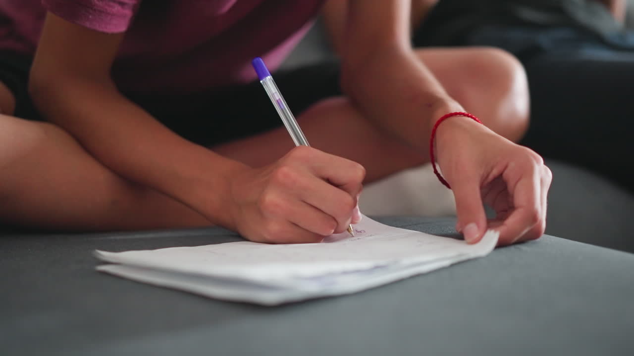 Niña pequeña sentada con las piernas cruzadas en el sofá escribiendo en un papel blanco con un bolígrafo azul, con una pulsera de cuerda roja en la muñeca, mientras que la pierna de otra persona aparece a su lado durante un momento de aprendizaje tranquilo y concentrado.