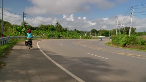 Mujer-Joven-Nómada-Montando-Una-Bicicleta-A-Través-Del-Arcén-De-La-Carretera-En-Un-País-Rural-Tropical