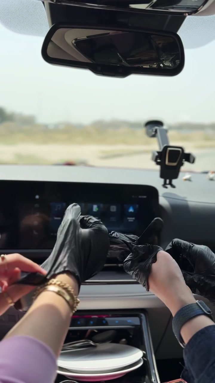 Primo piano di mani che indossano guanti monouso neri all'interno dell'abitacolo di un'auto durante il giorno, preparandosi per mangiare o pulire, con uno sfondo sfocato del cruscotto, in formato verticale per smartphone.