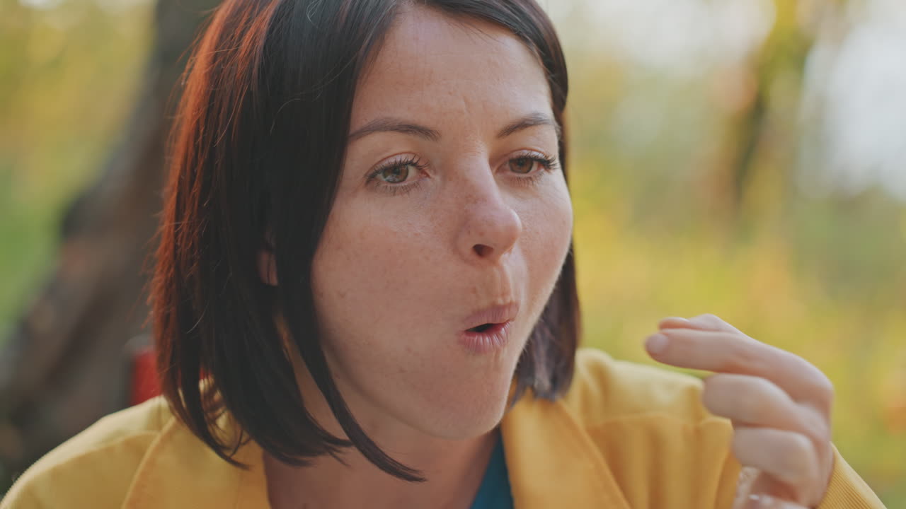 Primo piano di una donna in campeggio che assaggia uno spuntino, campeggiatrice contemplativa seduta nell'accampamento con giacca gialla, luce solare soffusa attraverso gli alberi, masticazione lenta e consapevole e sguardo pensieroso che cattura l'atmosfera tranquilla dell'aria aperta