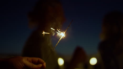 closeup hand holding burning sparkler. unknown people dancing at night party.