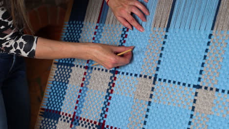 weaving pattern with blue and beige yarn on loom, hands adjusting with tool