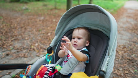 little toddler lying in the pram outdoors. baby boy starts to cry rubbing his eyes. nature blurred backdrop.