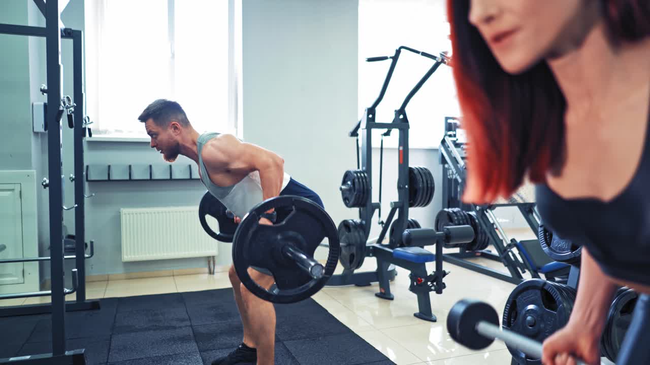 Muscular bearded man having workout together with beautiful woman. Healthy man and a woman training with heavy barbells in sports center.