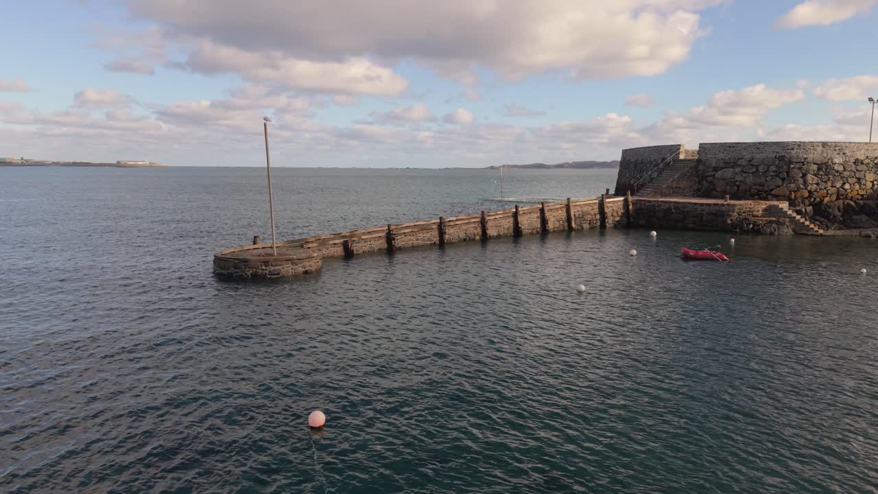 Riprese con drone a bassa quota a Salerie Corner, St Peter Port, Guernsey, che si concentrano sul molo di un antico porto a metà marea con una piccola barca all'ancora, con vista su Herm sotto il sole del tardo pomeriggio.