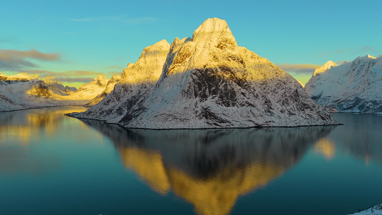 Et filmisk luftfoto af sneklædte tinder, der spejler sig i det rolige blå vand i en fjord i Lofoten, Norge, under en levende vintersolnedgang.