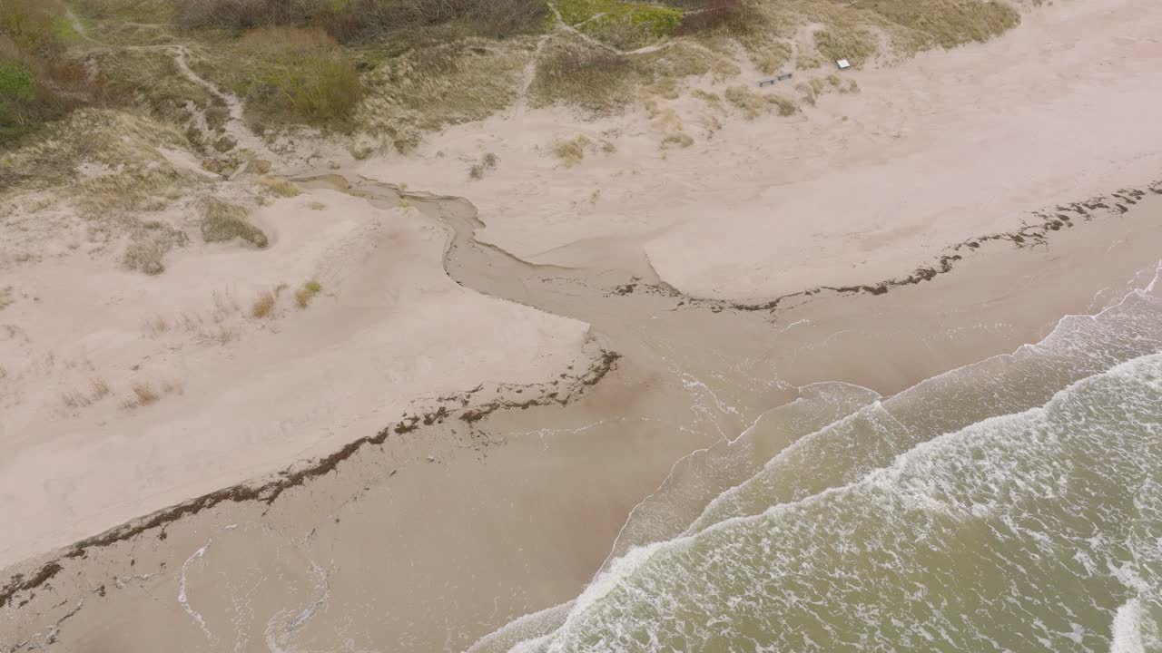 vista aérea de pájaro de la costa del mar báltico en un día nublado, playa de arena blanca, grandes olas de tormenta aplastando la costa, cambios climáticos, gran tiro de drones avanzando, inclinado hacia abajo