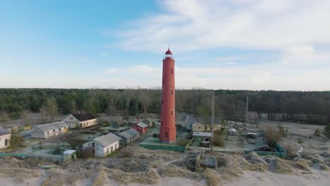 luftaufnahme des rot gefärbten leuchtturms von akmenrags, ostseeküste, lettland, weißer sandstrand, ruhige see, sonniger tag mit wolken, breite aufsteigende drohnenaufnahme, die sich vorwärts bewegt