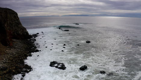 Grey-and-white-ocean-waves-roll-onto-black-stones-on-rocky-beach,-Aerial