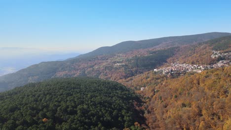 Vuelo-Con-Dron-Visualizando-La-Ladera-De-Una-Montaña-Con-Un-Pueblo-En-Un-Bosque-De-Castaños-Y-Pinos,-Girando-La-Vista-Hacia-La-Izquierda,-Dejando-Al-Fondo-Un-Valle-Con-Un-Cielo-Azul-En-Otoño-España