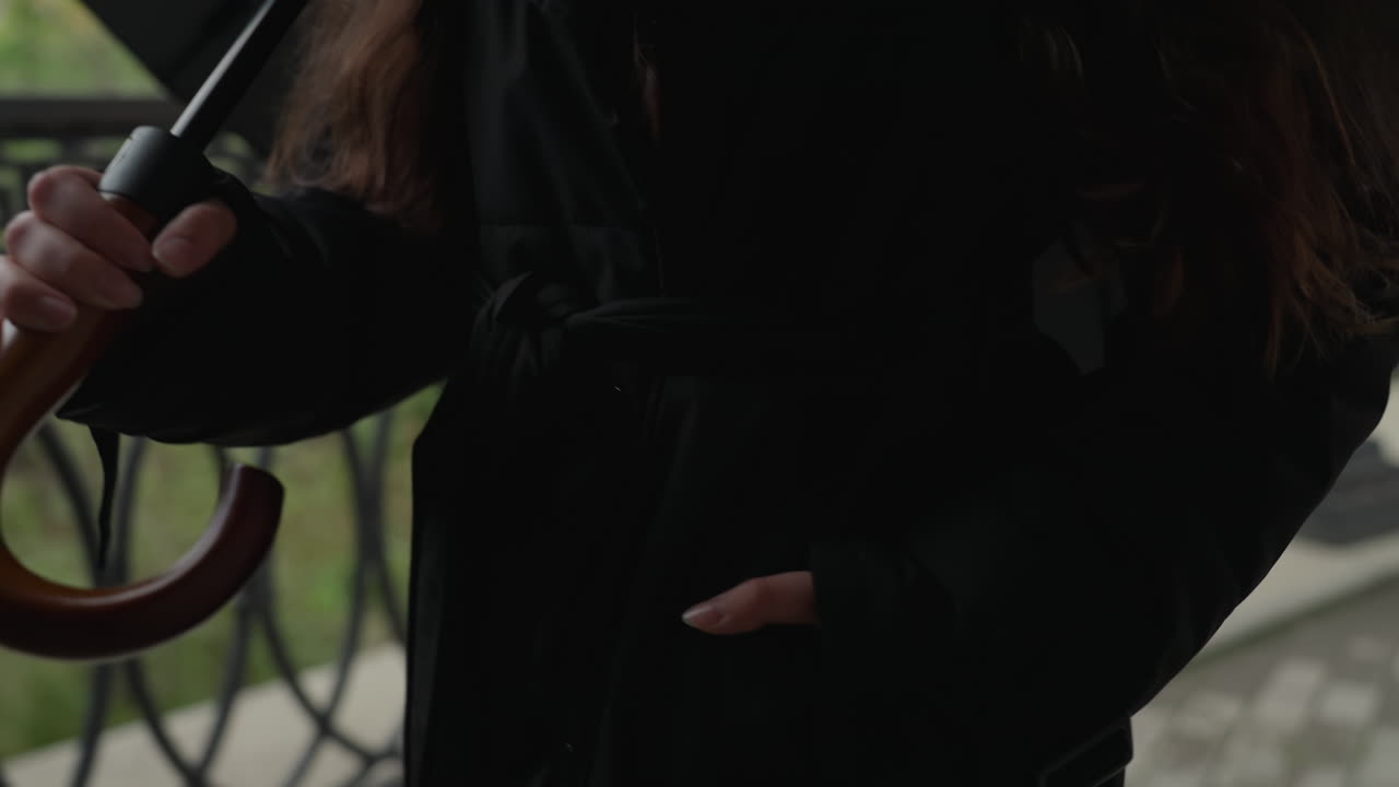 黒いコートを着て傘の柄を持ち、物思いにふける女性が、どんよりとした秋の散歩中に街のバルコニーに立っている。片方の手はポケットに、木の杖のグリップ、わずかな動き、物憂い雰囲気。