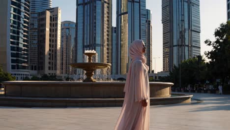 elegant woman wearing traditional clothing is walking near a fountain in a modern city square, surrounded by tall buildings and trees, enjoying the urban environment