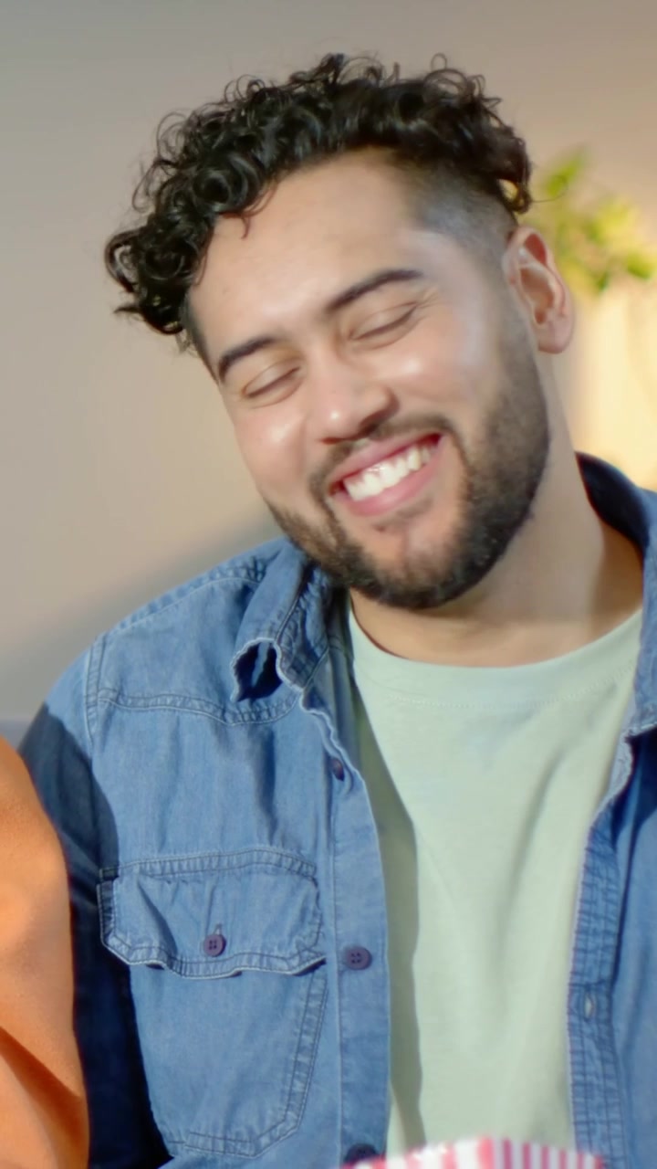Video verticale: Reagendo a una voce fuori campo, uomo in denim che si gira ridendo sul divano con i popcorn.