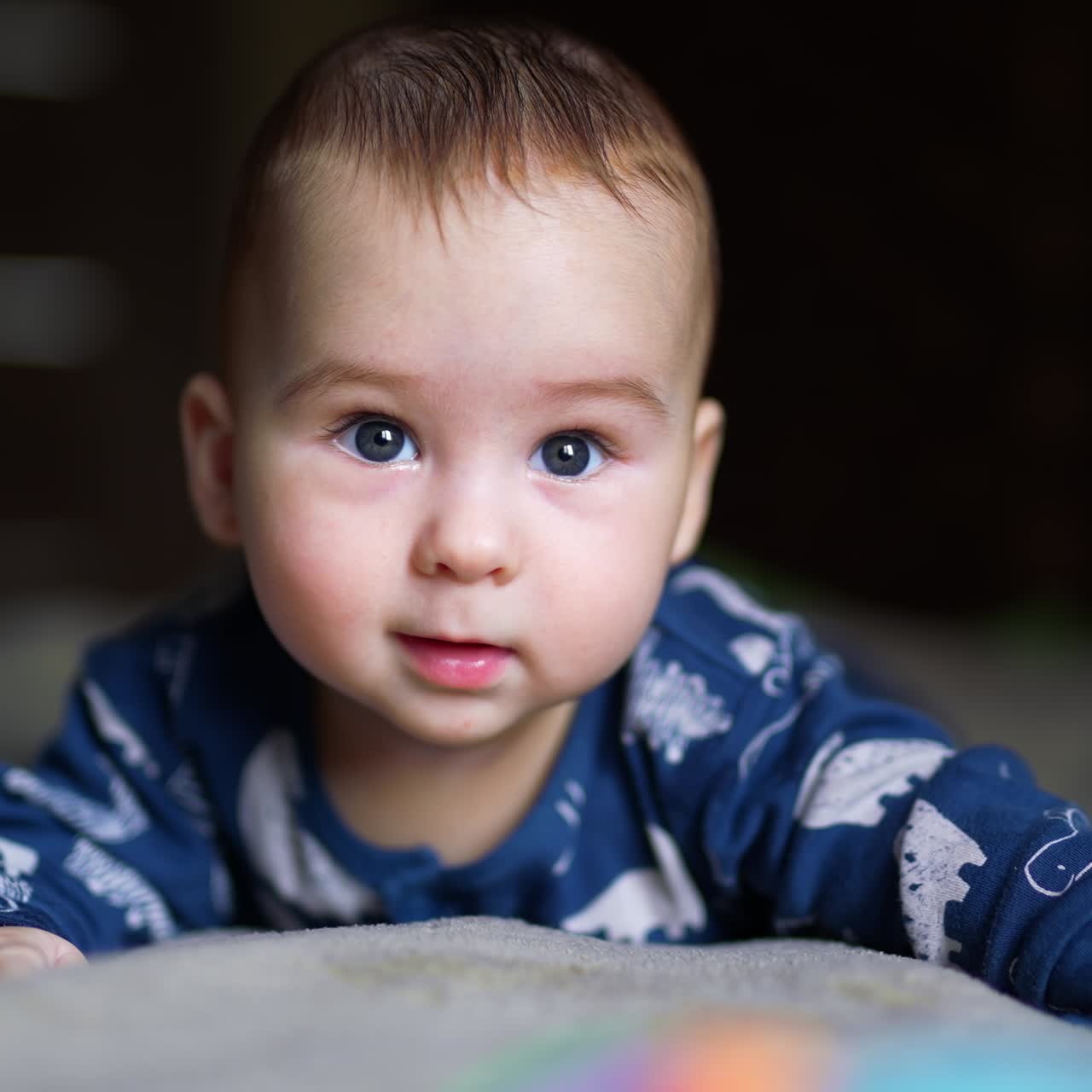 Cute half-year old boy lies peacefully on the bed. Adorable child smiles sweetly to the camera. Blurred backdrop