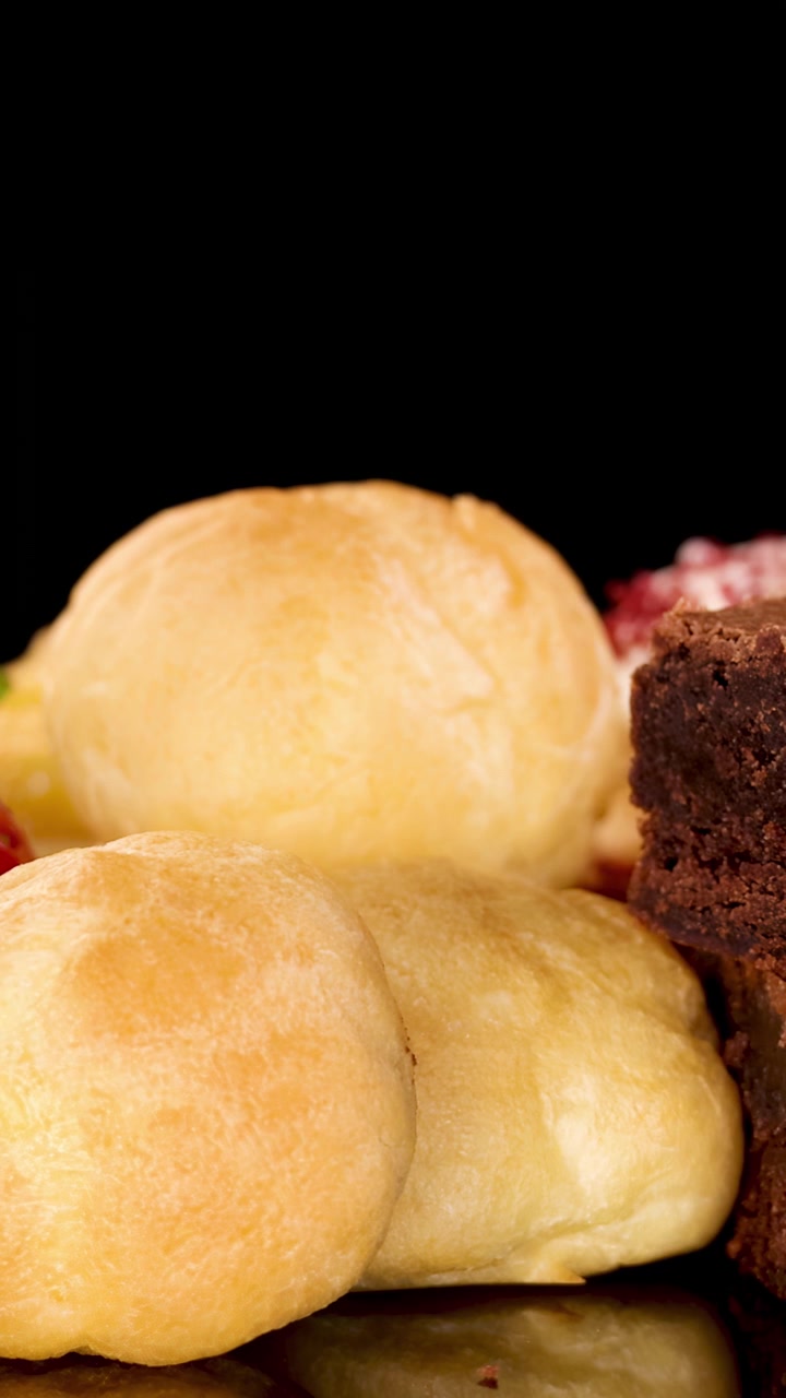A hand carefully stacks chocolate brownies next to cream puffs and fresh strawberries on a reflective black surface, under bright studio lighting with a close-up perspective