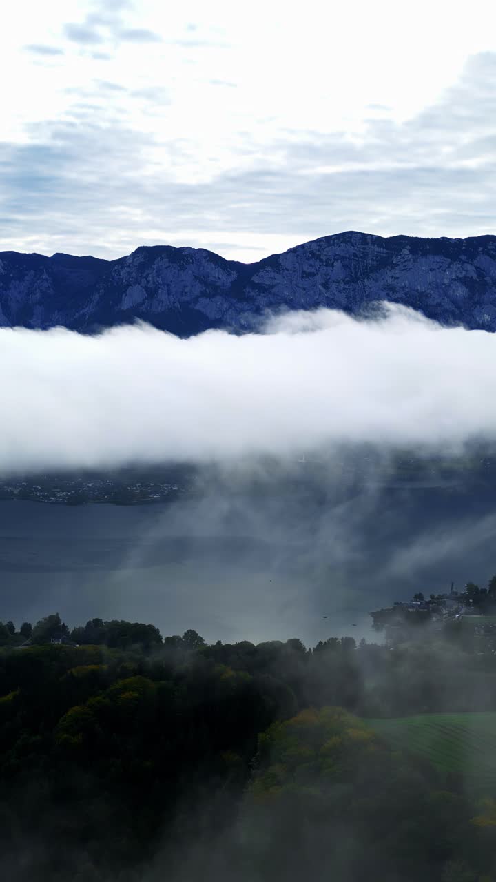 Clouds rolling over tall green mountains and valleys covered in morning fog descending to fjord water, aerial vertical