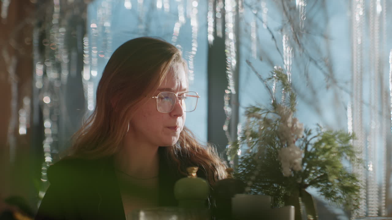 White woman in winter cafe sipping tea and planning, sunlight through window, icicles outside, plants and candles on table, glasses and blazer, quiet contemplative mood, gentle bokeh and warm tones