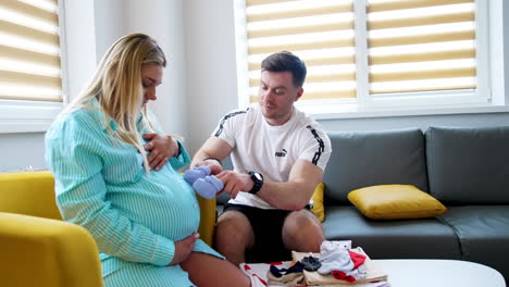 blonde woman with big belly sits on the sofa. her husband takes little baby socks and steps them gently by the abdomen. happy expecting couple in the hospital.