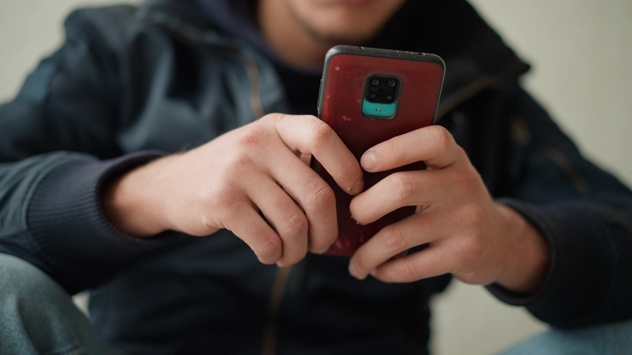 Close up of young man holding and interacting with red smartphone focusing on his hands gripping device. scene is indoors with soft lighting showing consistent engagement without significant