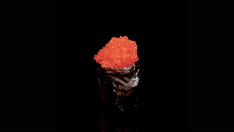a single piece of gunkan maki sushi topped with bright orange fish roe rotates smoothly against a seamless black background, under even studio lighting