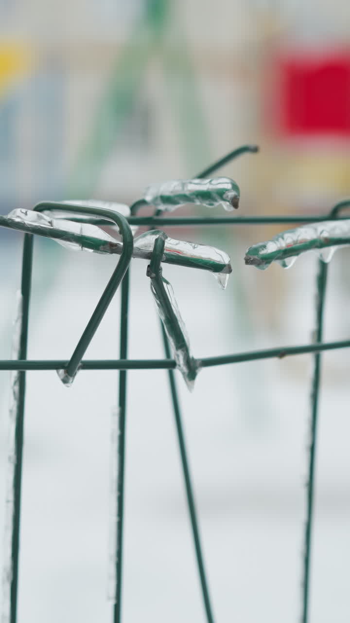 primer plano de una valla metálica verde cubierta de hielo con hielo colgando de los cables, en contra de un fondo nevado borroso y un paisaje invernal urbano