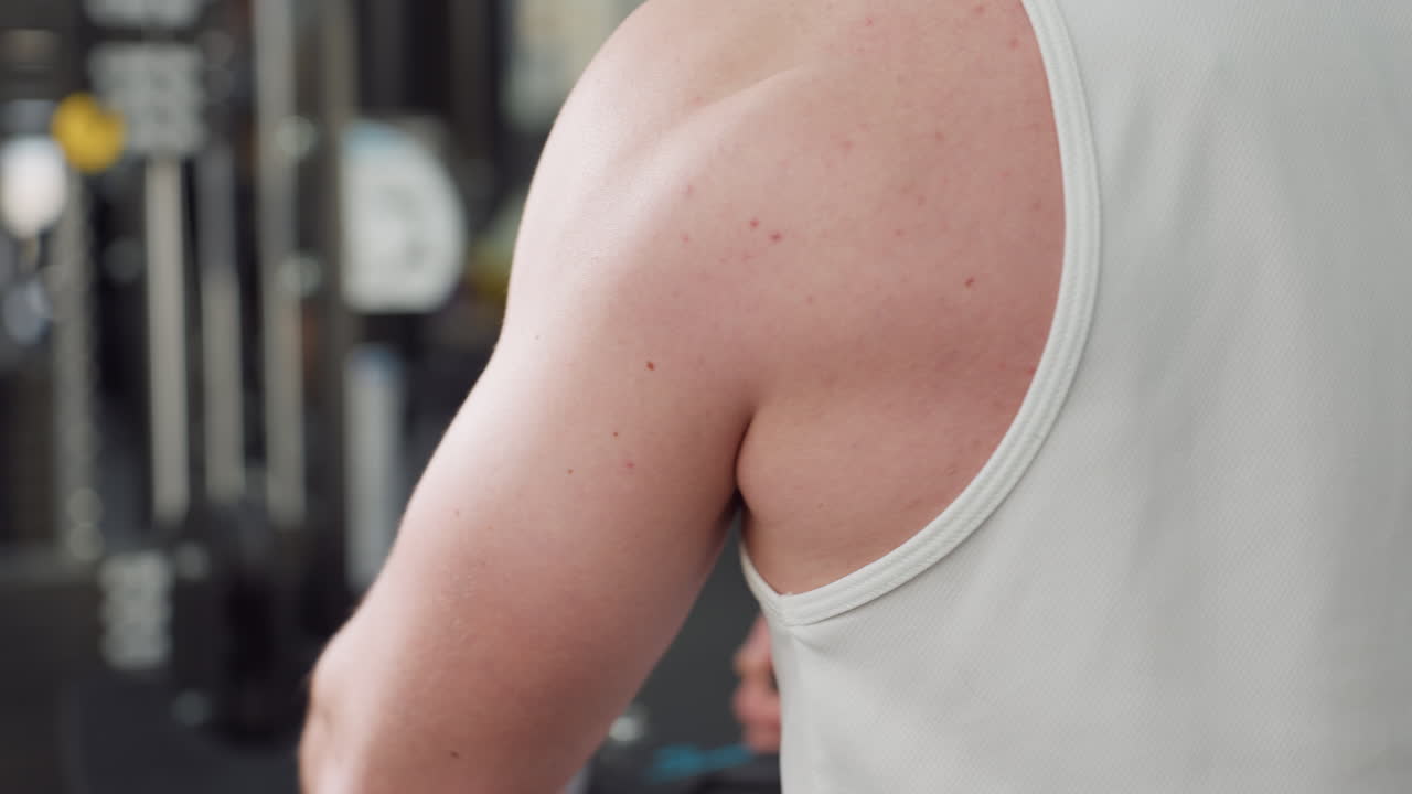 Shoulder view of man training on weight machine in gym center, muscles flexing under sleeveless shirt as he adjusts resistance and pushes handle with focused effort, showcasing strength and intensity
